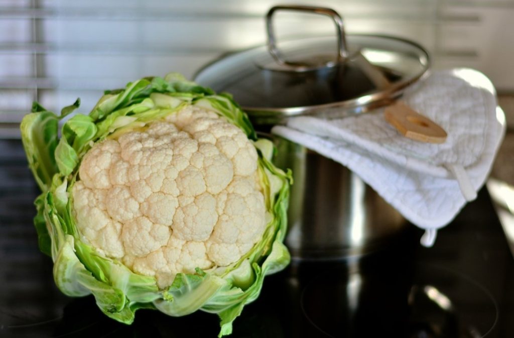 Cauliflower head with leaves attached next to a pot with a wooden spoon in it.