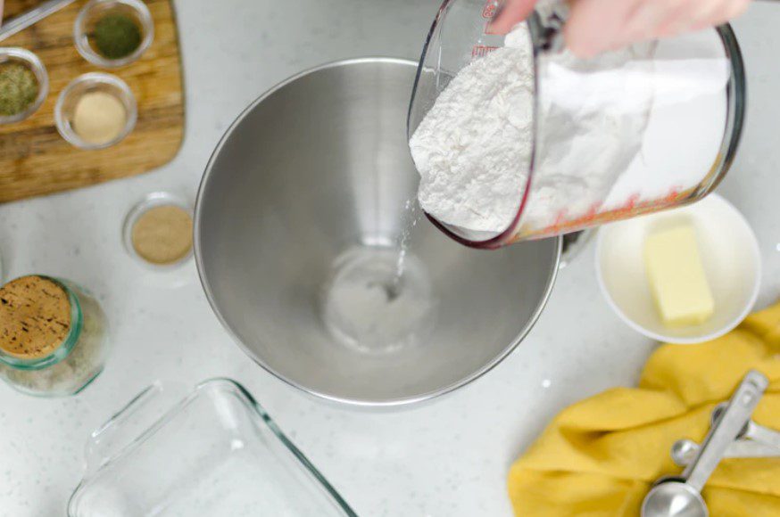 cake flour being poured into a metal bowl, butter and ingredients on table.