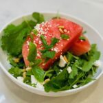 Large slice of watermelon on bed of arugula in white bowl with feta and basil.